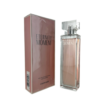 A bottle of Eternity Moment EDP by Calvin Klein with the cap off, next to its pink box packaging labeled 'ETERNITY MOMENT'.